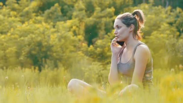 Glad ung kvinna som sitter på grön gräsmatta och prata på telefon eller Smartphone — Stockvideo