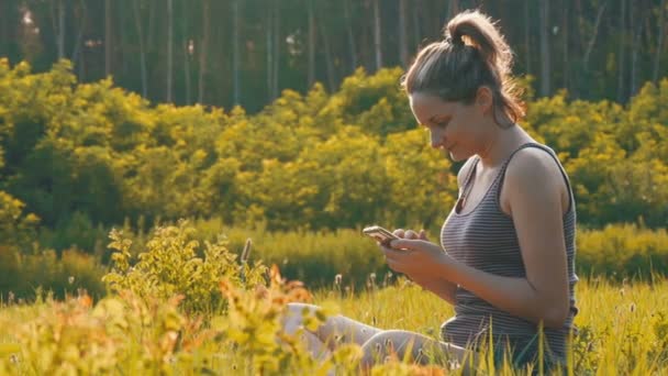 Mujer joven y feliz sentada en el césped verde y utiliza Smartphone en el campo escénico al atardecer — Vídeos de Stock