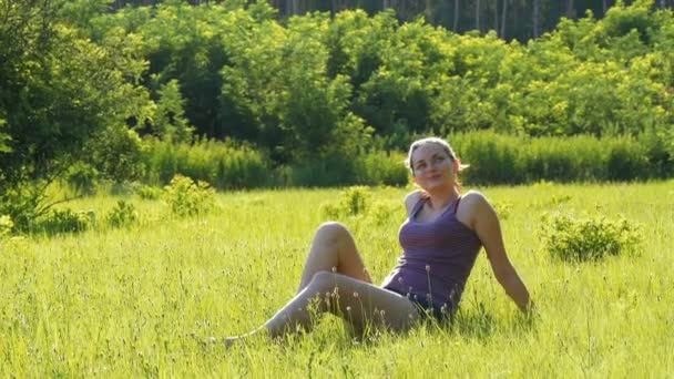 Glückliche junge Frau sitzt auf einem grünen Rasen in einem landschaftlich reizvollen Feld vor Sonnenuntergang. — Stockvideo