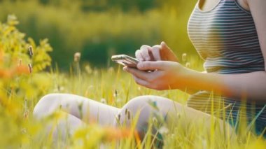 Yeşil çim ve doğal günbatımı arka plan sahasının kullanır Smartphone üzerinde oturan mutlu genç kadın