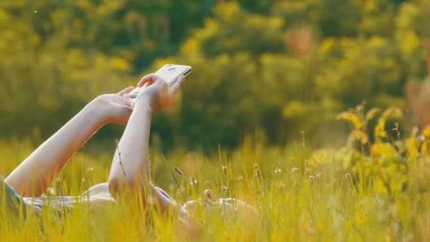 Mujer joven feliz acostada en el césped verde utiliza Smartphone en el campo escénico al atardecer Fondo — Vídeos de Stock