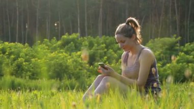Yeşil çim ve doğal günbatımı arka plan sahasının kullanır Smartphone üzerinde oturan mutlu genç kadın