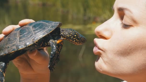 Женщина держит маленькую речную черепаху в руках у лица на фоне природы и реки — стоковое видео