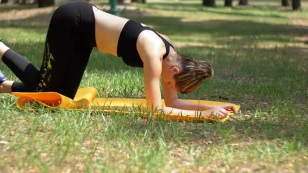 Joven atleta vestida con ropa deportiva practicando yoga acostada en una alfombra en un parque en un césped verde — Vídeos de Stock