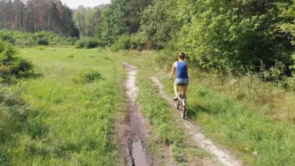 Vista aérea de la chica está montando una bicicleta retro en un camino de tierra en un campo cerca del bosque . — Vídeo de stock