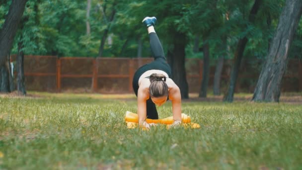 Joven atleta vestida con ropa deportiva practicando yoga acostada en una alfombra en un parque en un césped verde — Vídeo de stock