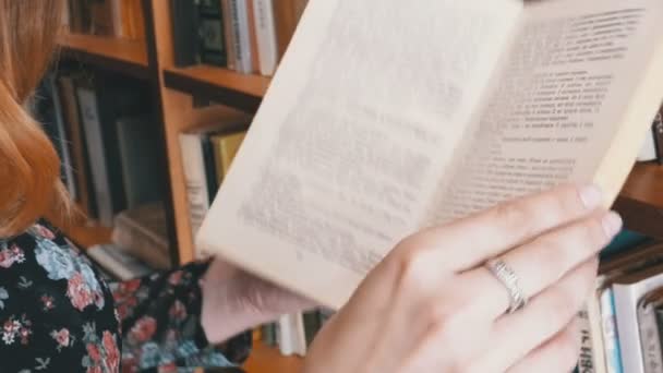 Lány a könyv tartja a kezében, és olvasson a könyvtárban, a háttérben a könyvespolcok — Stock videók