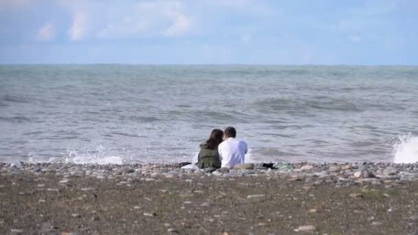 Пара мужчин и женщин сидят на каменистом пляже на фоне морского шторма — стоковое видео