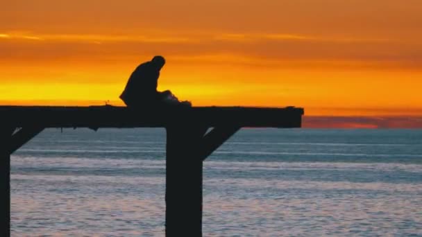 Silhueta de um homem solitário sentado no cais do mar contra o pôr do sol — Vídeo de Stock