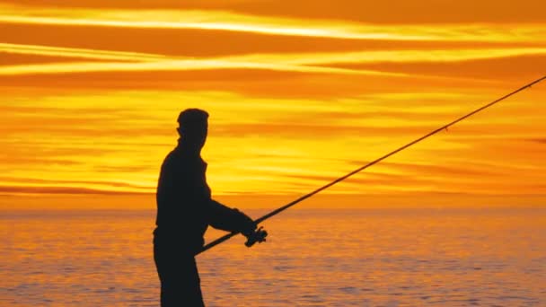 Silueta de un pescador con caña de pescar al atardecer sobre el mar — Vídeos de Stock