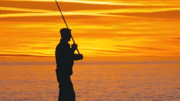 Silhueta de um pescador com uma cana de pesca ao pôr do sol sobre o mar — Vídeo de Stock