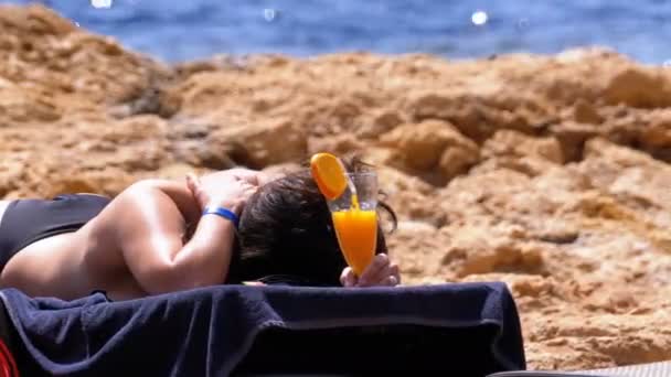 Красива дівчина лежить на вітальні на пляжі з коктейлем в склі в її руках — стокове відео