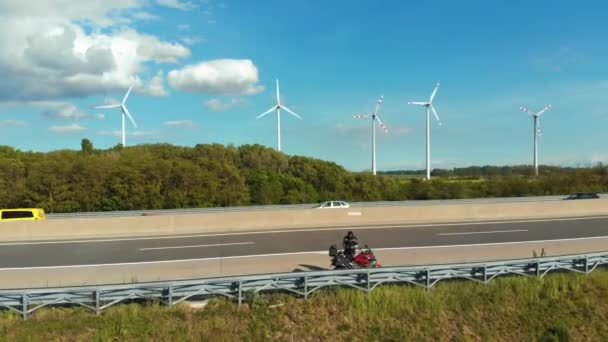 Otobanda motosikletin üzerinde duran yolcu, rüzgar çiftliklerinin yakınında bir insansız hava aracını havalandırdı. Avusturya — Stok video