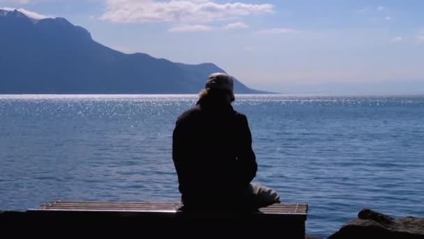 Silhouette di un uomo anziano e solitario seduto su una panchina sullo sfondo di un lago e montagne — Video Stock