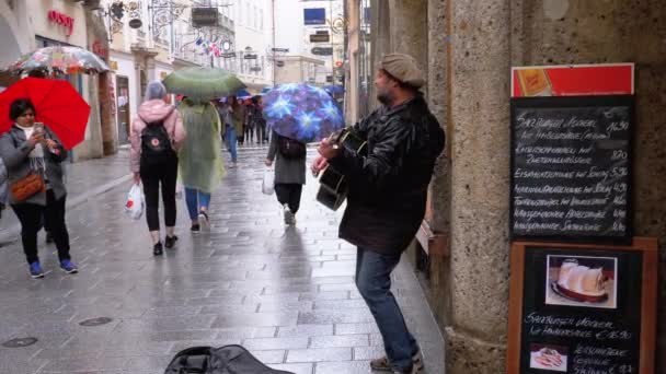 Muzyk uliczny grając na gitarze na słynnej ulicy w starym mieście w Salzburgu w deszczu. Austria — Wideo stockowe