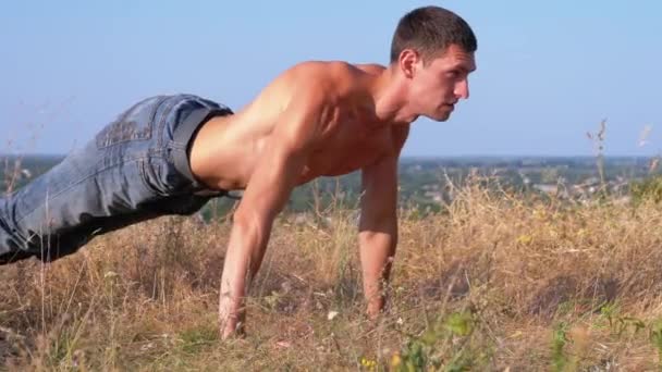 Jovem atlético com um tronco nu realiza flexões sobre a natureza — Vídeo de Stock