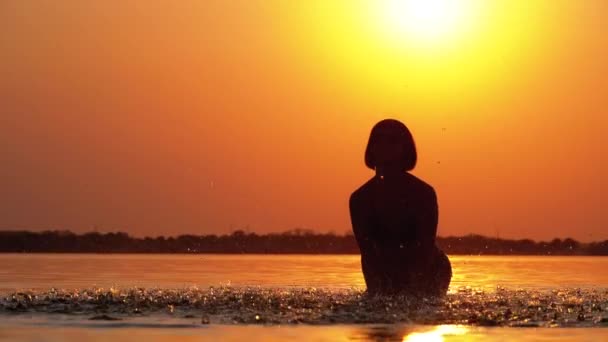 La silhouette de femme au coucher du soleil lève les mains et crée des éclaboussures d'eau. Mouvement lent — Video