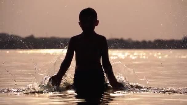 Силуэт Счастливого мальчика на закате, создающего брызги воды своими руками. Медленное движение — стоковое видео