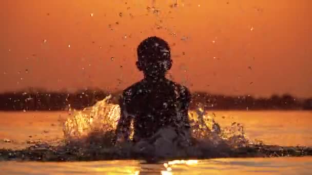 Σιλουέτα του αγοριού στο ηλιοβασίλεμα στο ποτάμι δημιουργώντας σταγόνες νερού με τα χέρια του. Αργή κίνηση — Αρχείο Βίντεο