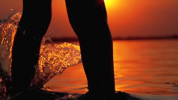 Σιλουέτα των ποδιών του αγοριού που τρέχει στο νερό στο ηλιοβασίλεμα και δημιουργώντας πιτσιλίσματα. Αργή κίνηση — Αρχείο Βίντεο