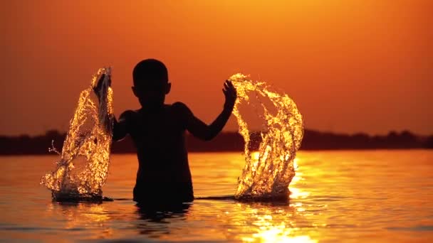 Silhouette de garçon au coucher du soleil lève les mains et créer des éclaboussures d'eau. Mouvement lent — Video