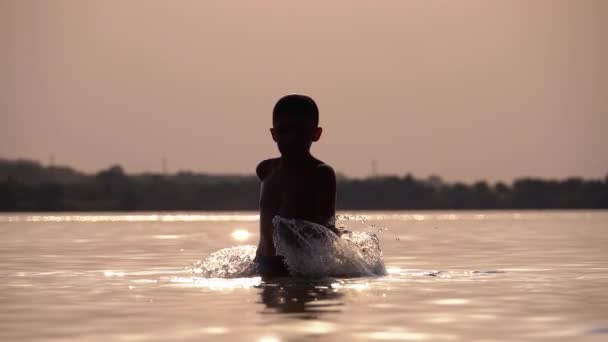 Silueta de Happy Boy al atardecer creando salpicaduras de agua con sus manos. Movimiento lento — Vídeo de stock