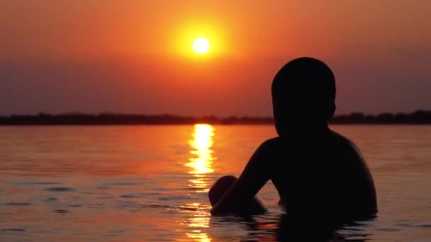 在日落和橙色路径的背景中坐在水中的男孩的剪影。慢动作 — 图库视频影像