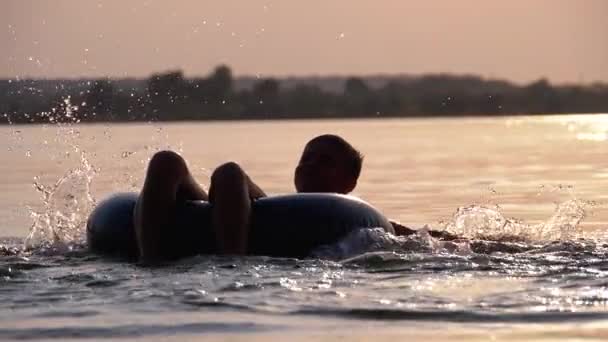 Silhuett av Happy Boy swiming på uppblåsbar cirkel i floden vid solnedgången. Slow motion — Stockvideo
