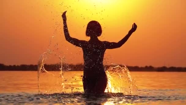 Silueta de mujer al atardecer levanta las manos y crea salpicaduras de agua. Movimiento lento — Vídeo de stock