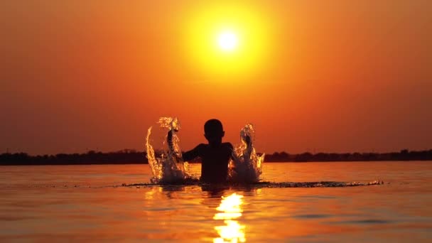 Silhuett av pojke vid solnedgången väcker händer och skapar stänk av vatten. Slow motion — Stockvideo