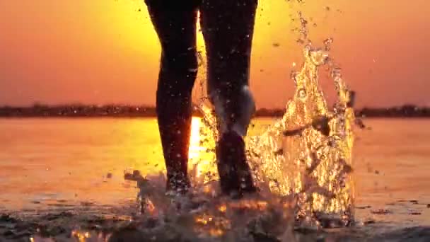 Silhouette der Beine eines Jungen, der bei Sonnenuntergang ins Wasser läuft und Spritzer erzeugt. Zeitlupe — Stockvideo