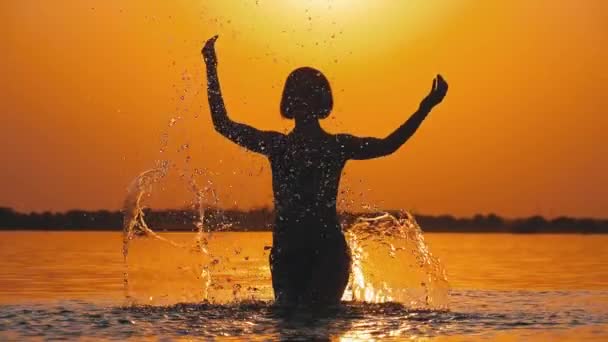 A silhueta da mulher no pôr-do-sol levanta as mãos e cria salpicos de água. Movimento lento — Vídeo de Stock