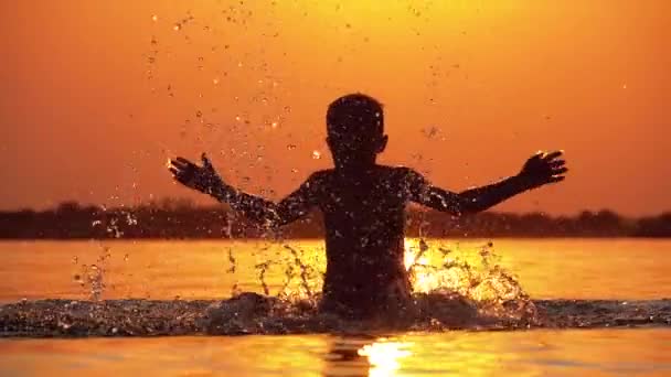 Silhouet van de jongen bij zonsondergang verhoogt de handen en het creëren van spatten van water. Slow Motion — Stockvideo