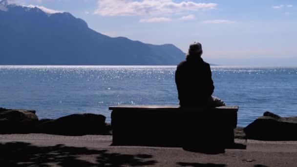 Silhouette of a Lonely Aged Man Sitting on a Bench on backdrop of a Lake and Mountains — Stock Video