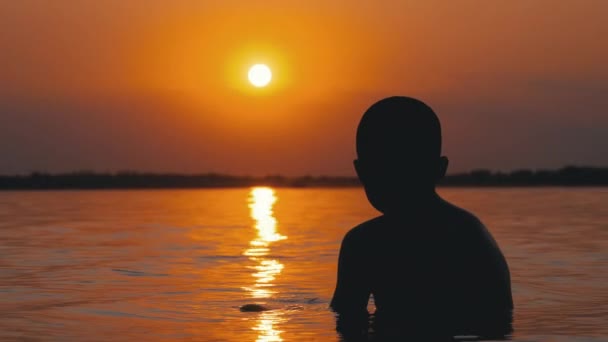 Silhouette d'un garçon assis dans l'eau sur fond de coucher de soleil et de sentier orange. Mouvement lent — Video