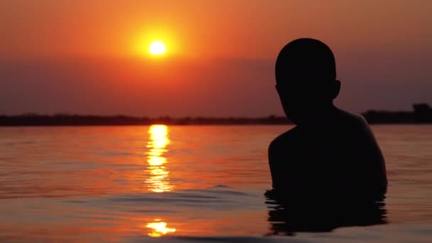 Silhouette of Boy Sitting in the Water on Background of Sunset and Orange Path (en inglés). Moción lenta — Vídeos de Stock