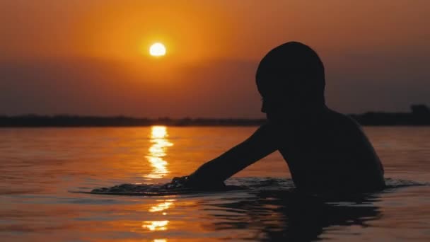 Silhouette eines Jungen, der im Wasser vor dem Hintergrund von Sonnenuntergang und orangefarbenem Pfad sitzt. Zeitlupe — Stockvideo