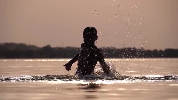 Silueta de Happy Boy al atardecer creando salpicaduras de agua con sus manos. Movimiento lento — Vídeo de stock