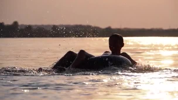 Силует щасливий хлопчик Swiming на надувному колі в річці на заході сонця. Повільний рух — стокове відео