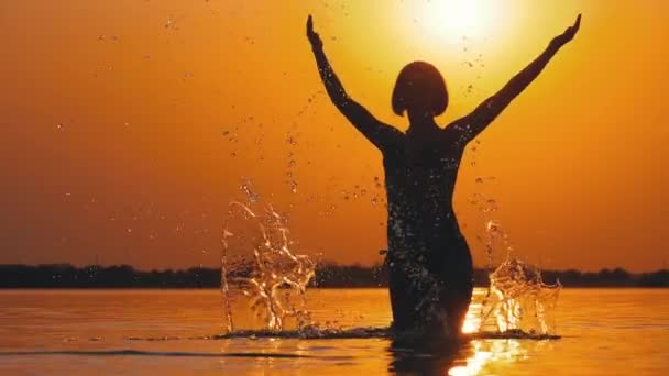 Σιλουέτα της γυναίκας στο ηλιοβασίλεμα ανεβάζει τα χέρια και δημιουργώντας σταγόνες νερού. Αργή κίνηση — Αρχείο Βίντεο