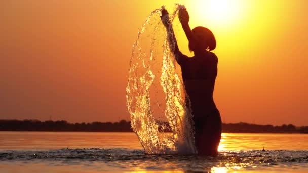 Силует жінки на заході піднімає руки вгору і створює бризки води. Повільний рух — стокове відео