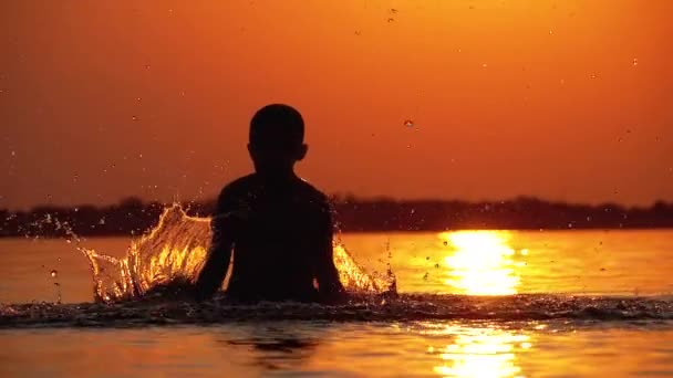 Silhouette of Boy al atardecer levanta las manos y crea salpicaduras de agua. Movimiento lento — Vídeos de Stock