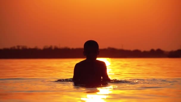 Silhuett av pojke vid solnedgången väcker händer och skapar stänk av vatten. Slow motion — Stockvideo