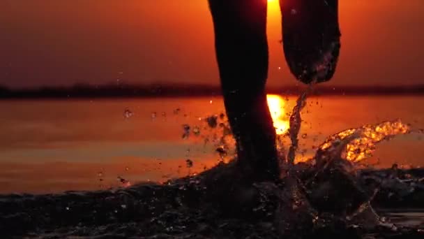 Silhuett av ben av pojke som rinner i vattnet vid solnedgången och skapa stänk. Slow motion — Stockvideo