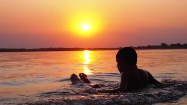 Silhouette d'un garçon assis dans l'eau sur fond de coucher de soleil et de sentier orange. Mouvement lent — Video