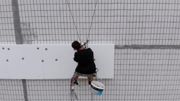 Воздушный вид на промышленный альпинист, подвешенный на канатах, выполняет работы по изоляции фасада высотного здания с использованием пенопласта — стоковое видео