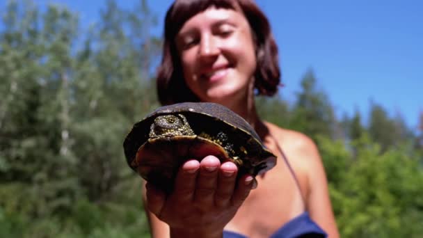 Женщина держит смешную черепаху в руках и улыбается на реке с зеленой растительностью — стоковое видео