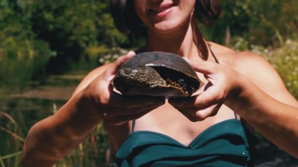 Женщина держит смешную черепаху в руках и улыбается на реке с зеленой растительностью — стоковое видео