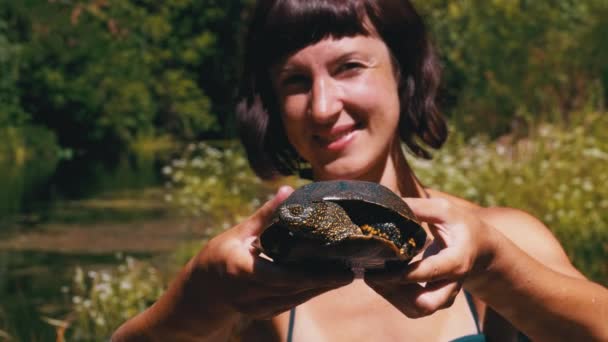 Mulher segura tartaruga engraçada no braço e sorri no rio com vegetação verde — Vídeo de Stock