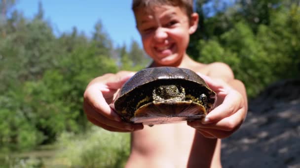 Menino segura tartaruga em braços e sorrisos Viciosamente no rio com vegetação verde — Vídeo de Stock